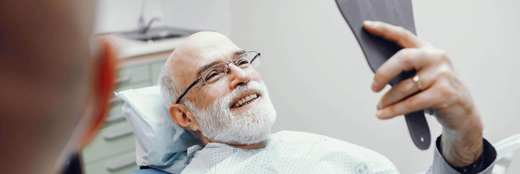 man smiling in dental hand mirror after getting crowns in barrie